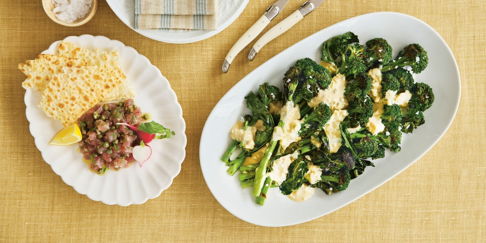 Photograph of a fresh dish of tuna tartare and Sardinian crackers on their left, a larger plate of barbequed broccoli with creamy anchovy sauce 
